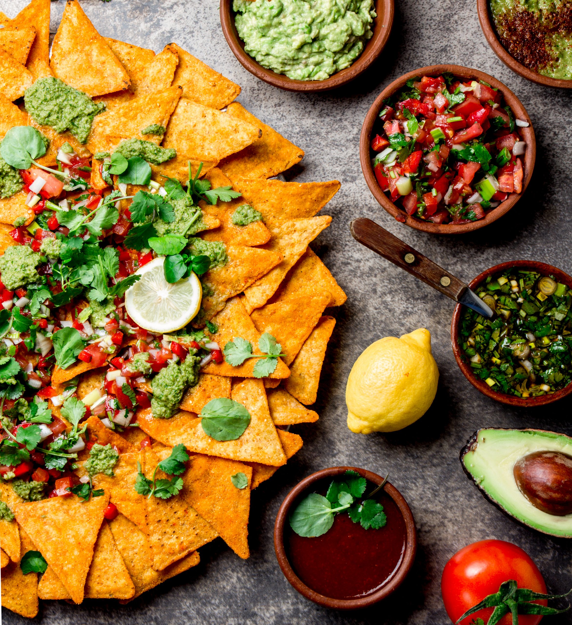 Nachos. Totopos with sauces. Mexican food concept. Yellow corn totopos chips with different sauces salsas – pico del gallo, guacamole, salsa verde, chili pebre and fresh avocado, tomatoes, lemon and cilantro on stone gray background, top view